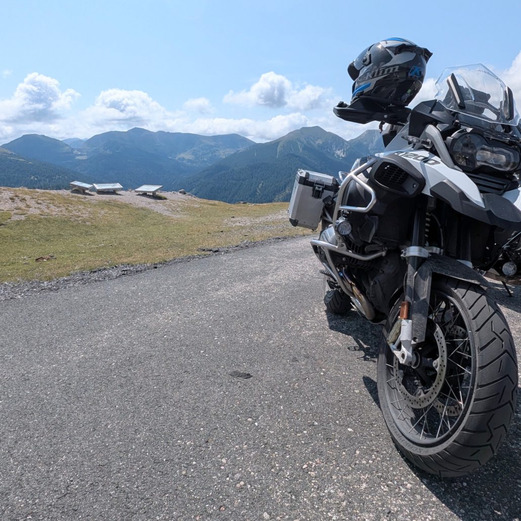 Weiße BMW R 1200 GS mit Alukoffern und Klim-Helm parkt auf Passstraße vor weitem Bergpanorama.