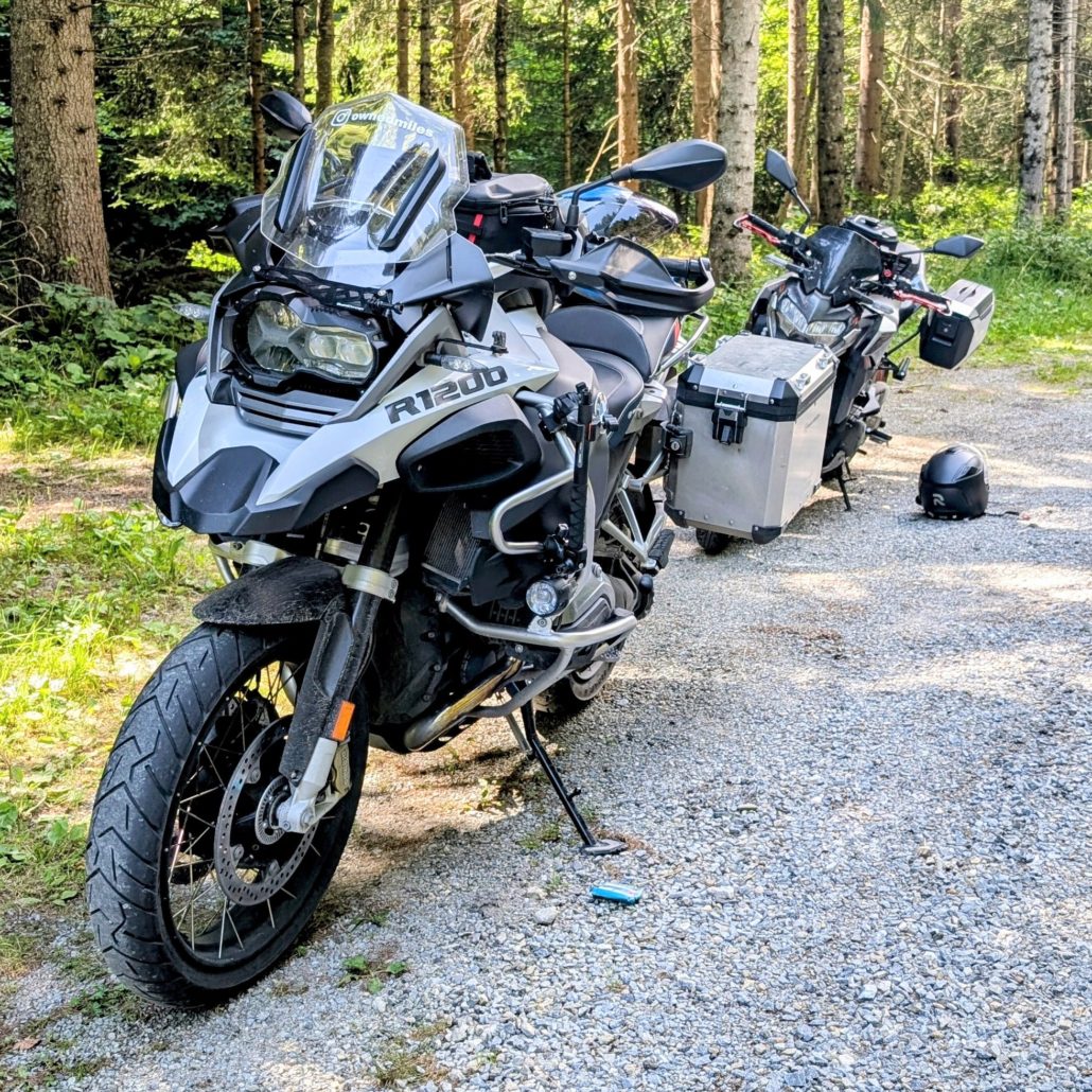 Zwei Reise-Enduros stehen auf einem Schotterweg in einem Wald. Vorne eine silberne BMW R 1200 GS mit Seitenkoffern.