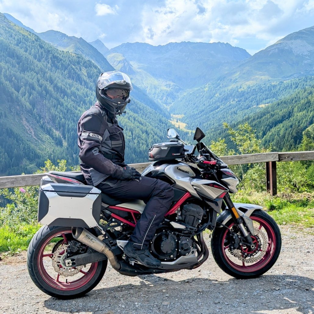 Motorradfahrer auf einer silbernen Kawasaki Z900RS mit Gepäck und roten Felgen an einem Aussichtspunkt in den Bergen.
