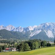 Ein sonniger Blick auf die Steinernes Meer Bergkette, davor eine grüne Wiese, Wälder und vereinzelte Häuser in einem Tal.