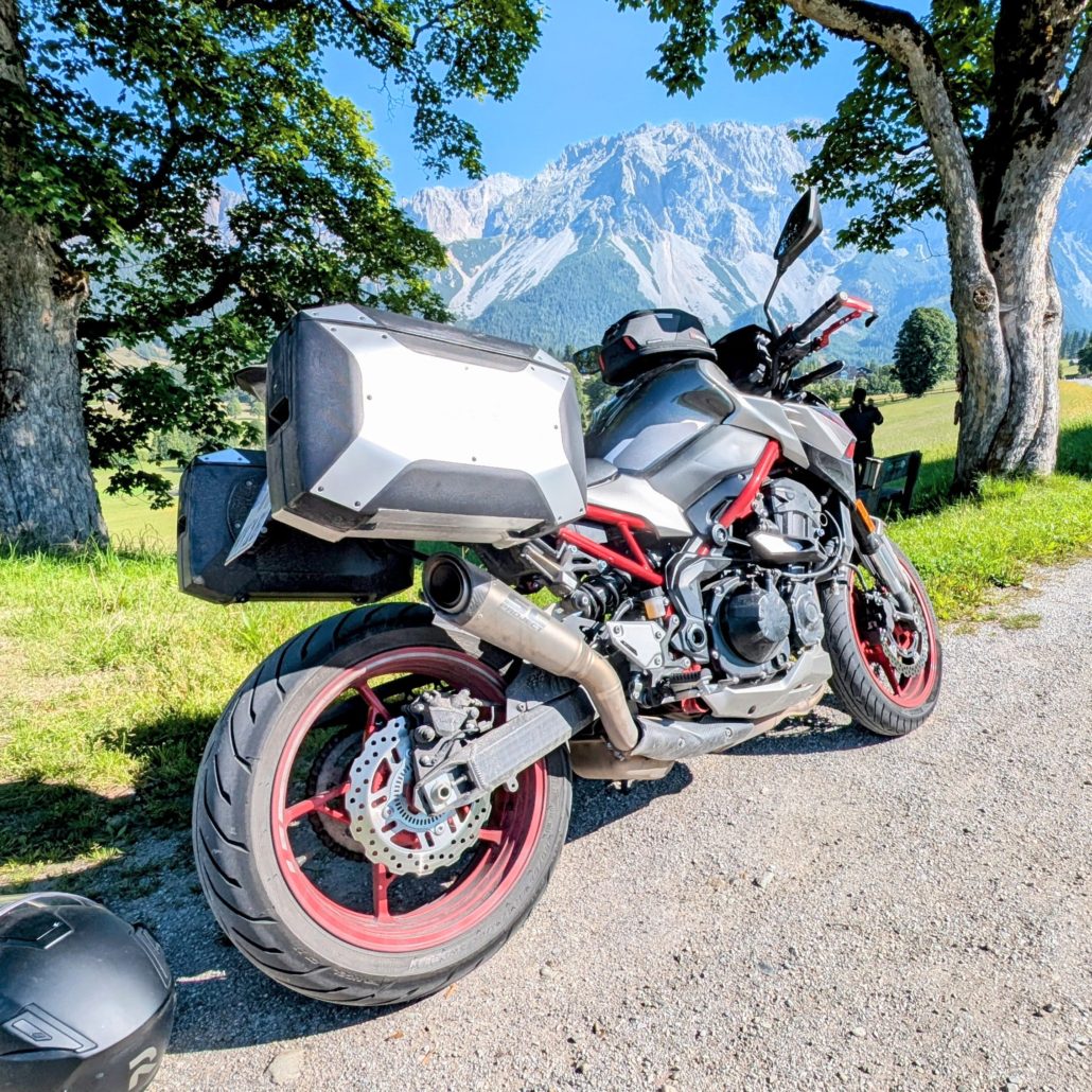 Graues Naked Bike (Kawasaki Z900) mit rotem Rahmen und Alu-Koffer auf Schotterweg vor einem Alpenpanorama geparkt.