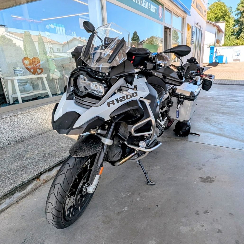 Weiße BMW R1200 GS Reiseenduro mit Sturzbügeln und Windschild vor einem Ladengeschäft geparkt.