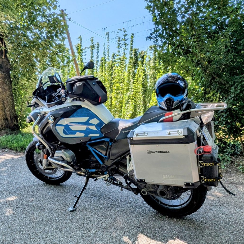 Weiß-blaues BMW R 1250 GS Motorrad mit silbernen Aluminiumkoffern und blauem Helm auf der Sitzbank. Instagram-Handle "@ownedmiles".