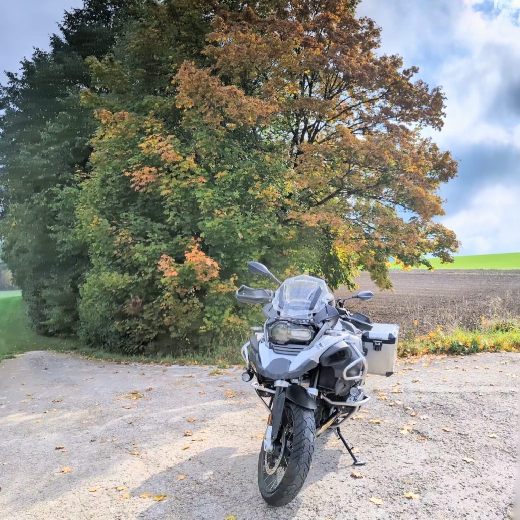 Graues BMW GS Motorrad mit Koffern steht auf einem Feldweg vor einem großen Baum mit Herbstlaub.