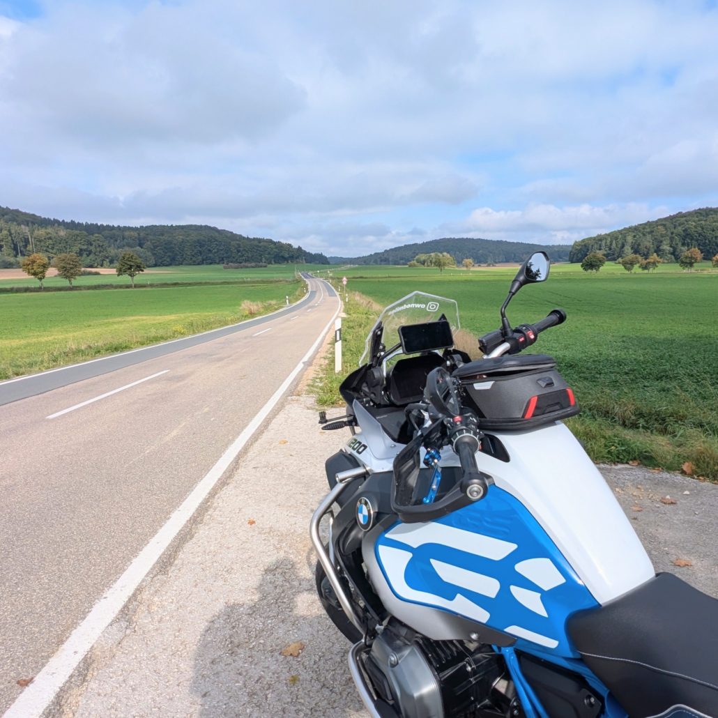 BMW R 1200 GS Motorrad mit blau-weißer Lackierung am Straßenrand, Blick auf eine Landstraße durch grüne Felder.