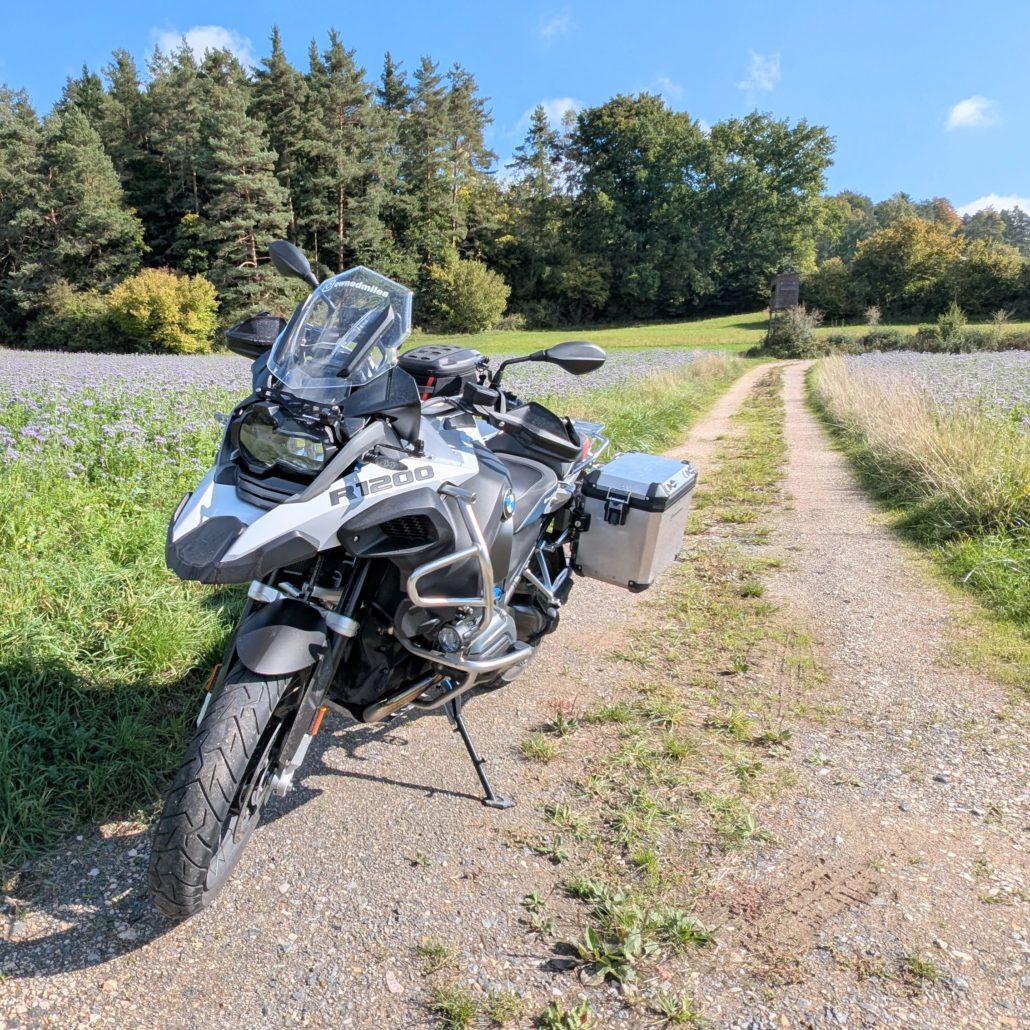 Weiße BMW R 1200 GS Adventure mit silbernen Koffern auf einem Schotterweg, umgeben von blühenden Feldern und Wald.