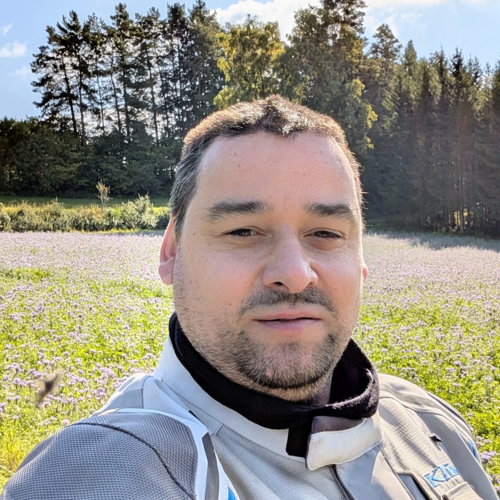 Mann in grauer Motorradjacke vor blühendem Phazelia-Feld und Wald unter blauem Himmel.