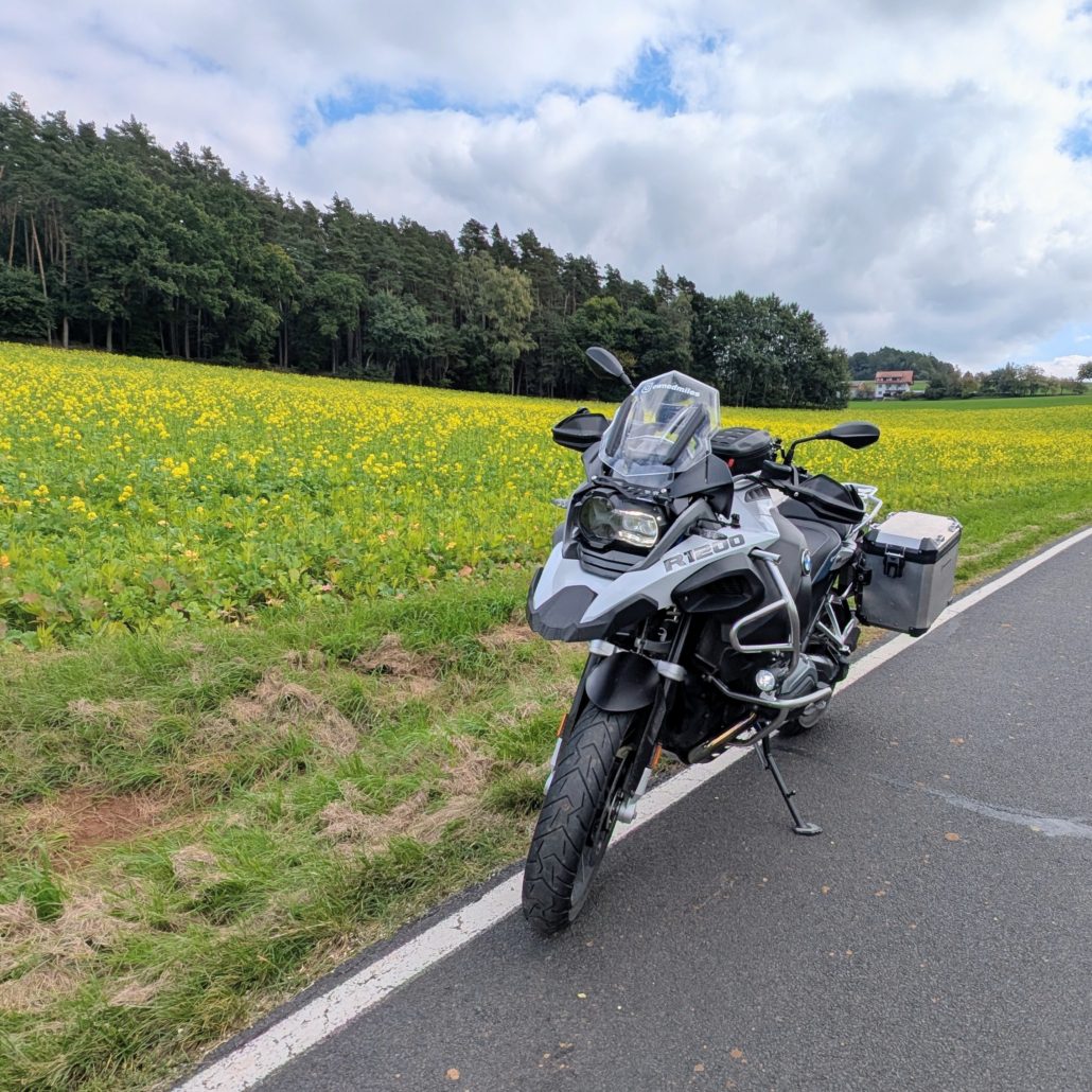 Ein silbergraues BMW R 1200 Motorrad steht auf einer Landstraße neben einem gelben Rapsfeld unter bewölktem Himmel.