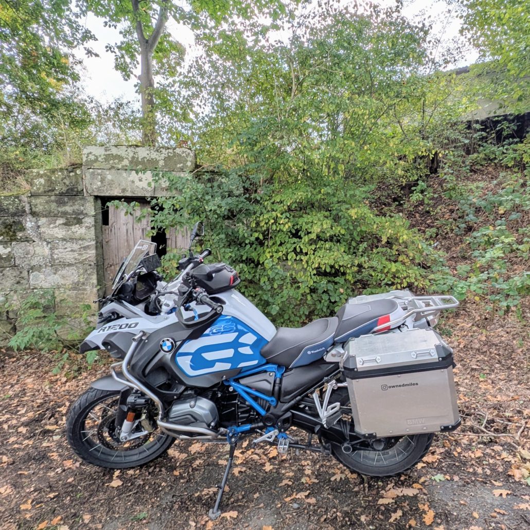 Geparktes weiß-blaues BMW R1200 Motorrad mit Alukoffern und Text "ownedmiles" vor einer zugewachsenen Steinmauer.