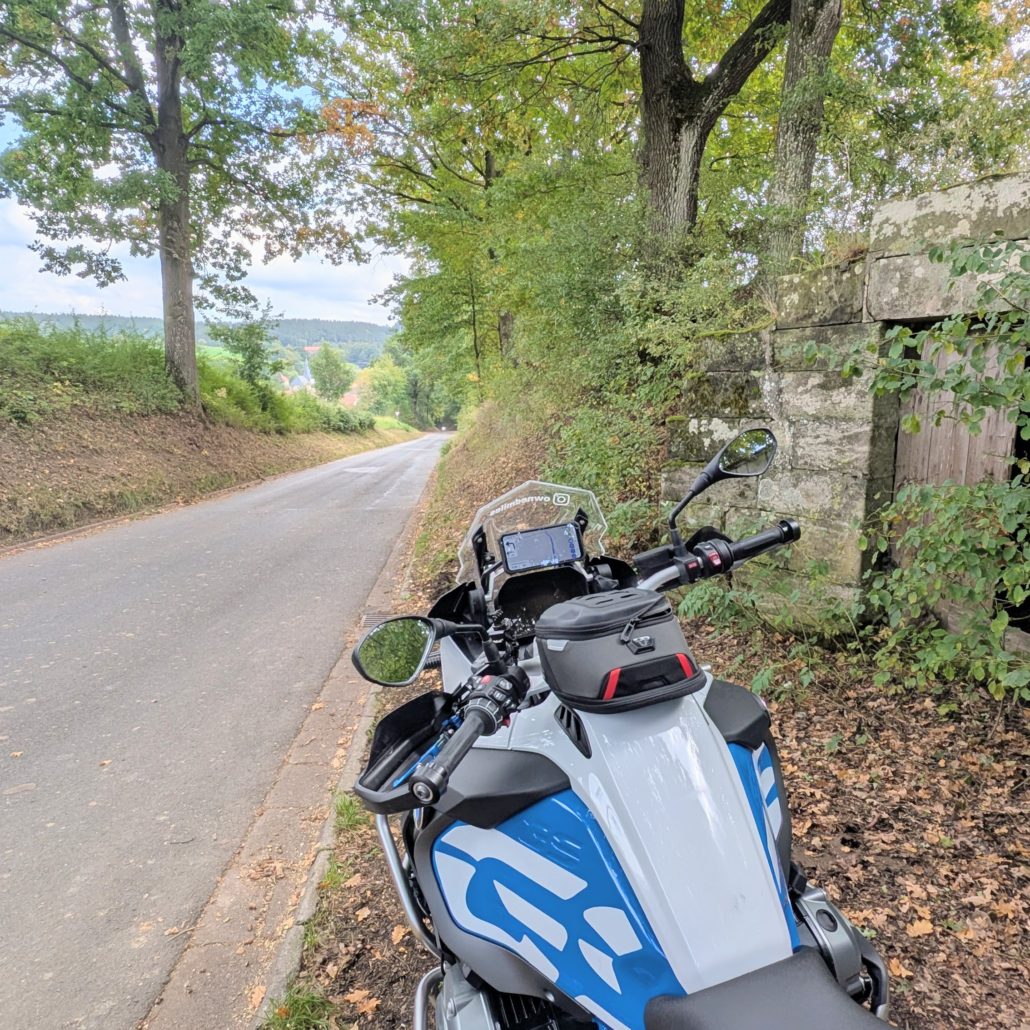 Auf einem Landstraßenrand parkendes, weiß-blaues BMW Motorrad mit Tankrucksack vor einer Steinmauer und Bäumen.