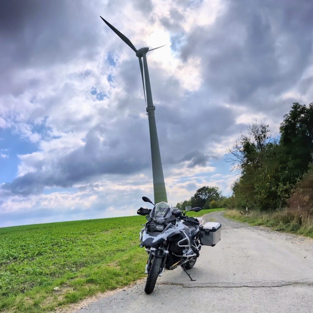 Die grau-weiße BMW R 1200 GS Adventure steht geparkt auf einem Feldweg neben einem Windrad und einer grünen Wiese.