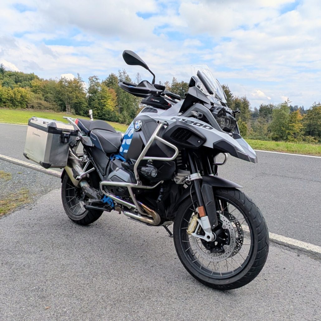 Grau-schwarze BMW R 1200 GS Adventure mit silbernen Seitenkoffern, auf einer Landstraße am Waldrand parkend.