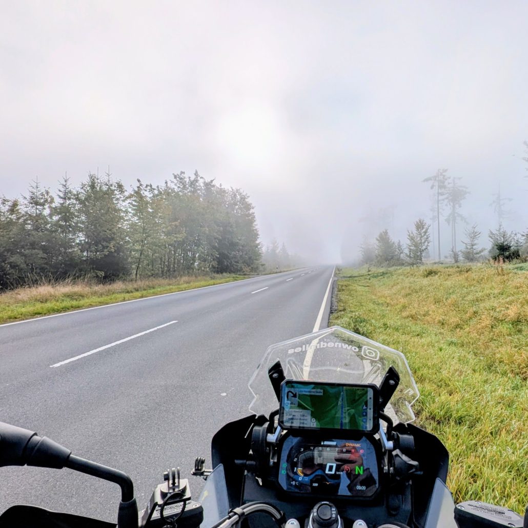 Fahrerperspektive auf eine Motorradarmatur mit TFT-Display und Navigations-Smartphone auf einer leeren, nebligen Landstraße.