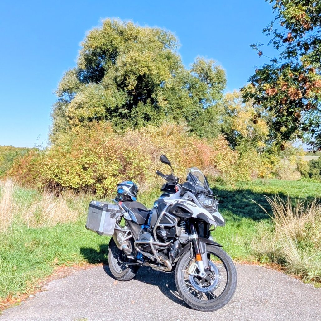 Weiß-schwarze BMW R 1200 GS Adventure mit silbernen Seitenkoffern, geparkt auf Asphaltweg vor Herbstlaub.