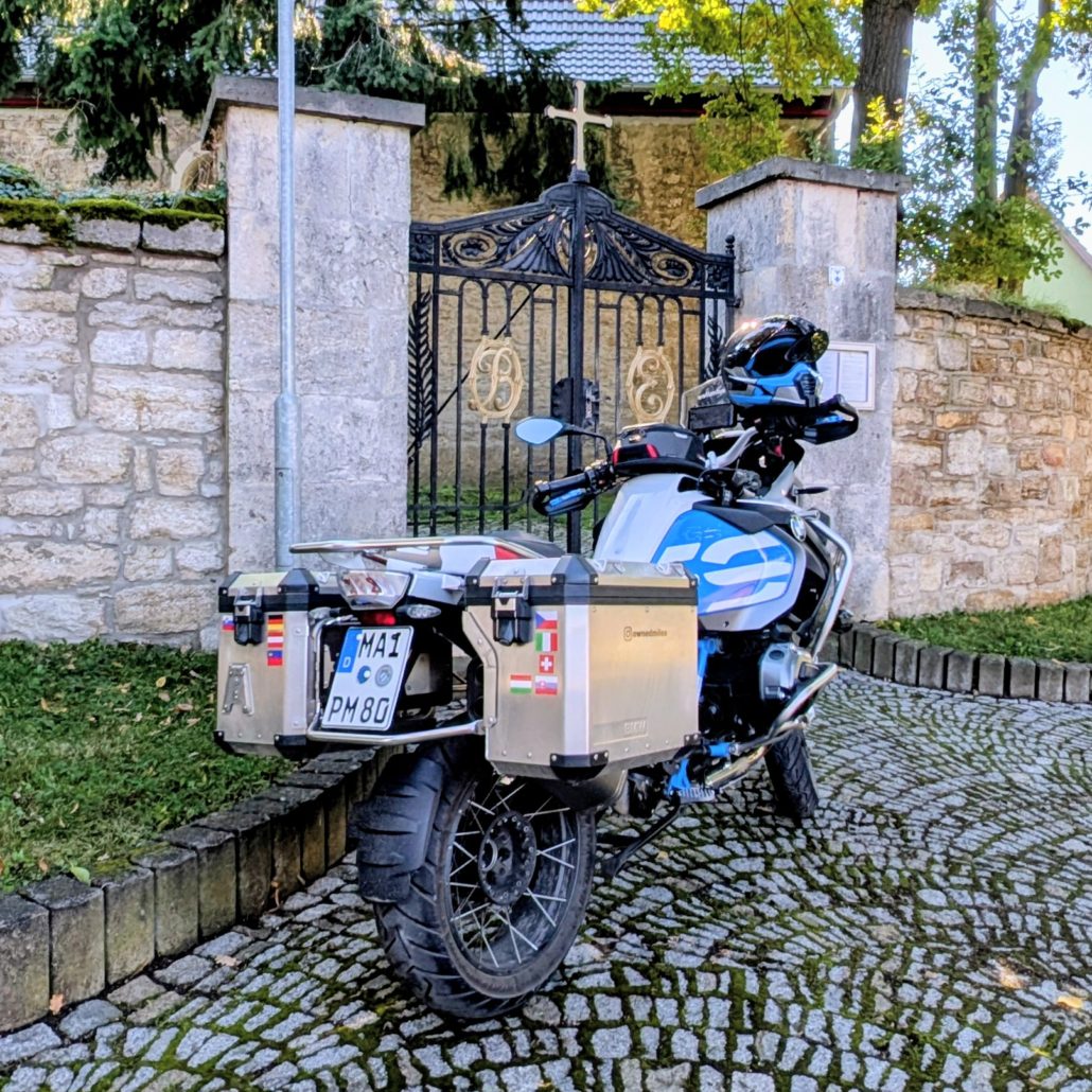 BMW R 1200 GS Adventure Motorrad, weiß-blau, mit Aluminiumkoffern vor einem kunstvoll geschmiedeten Friedhofstor.
