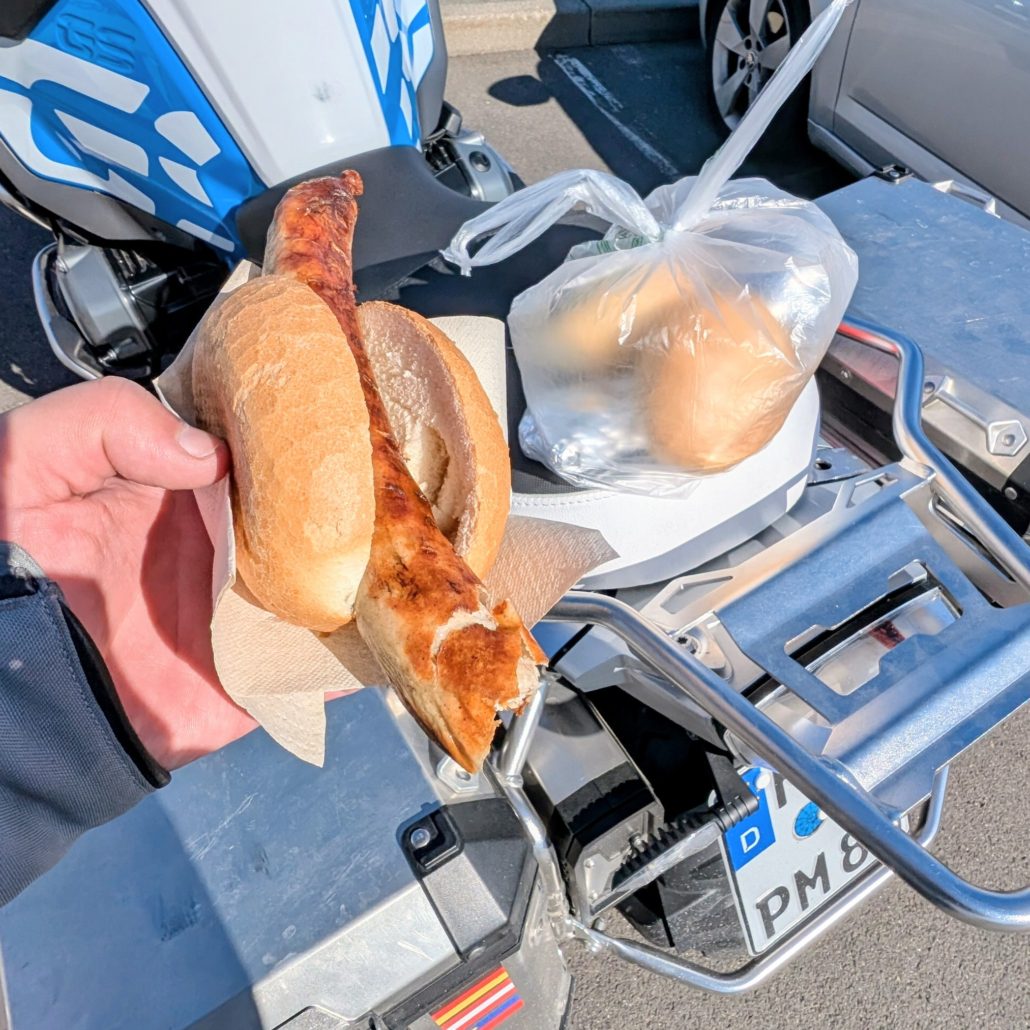 Geröstete Bratwurst in einer Semmel, gehalten von einer Hand über dem silbernen Gepäckträger eines Motorrads.