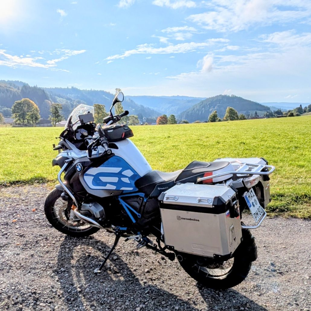 BMW R 1250 GS Adventure Motorrad mit weißen und blauen Akzenten, Aluminium-Koffern ("ownadmiles") auf Schotterweg vor einer sonnigen, grünen Hügellandschaft.