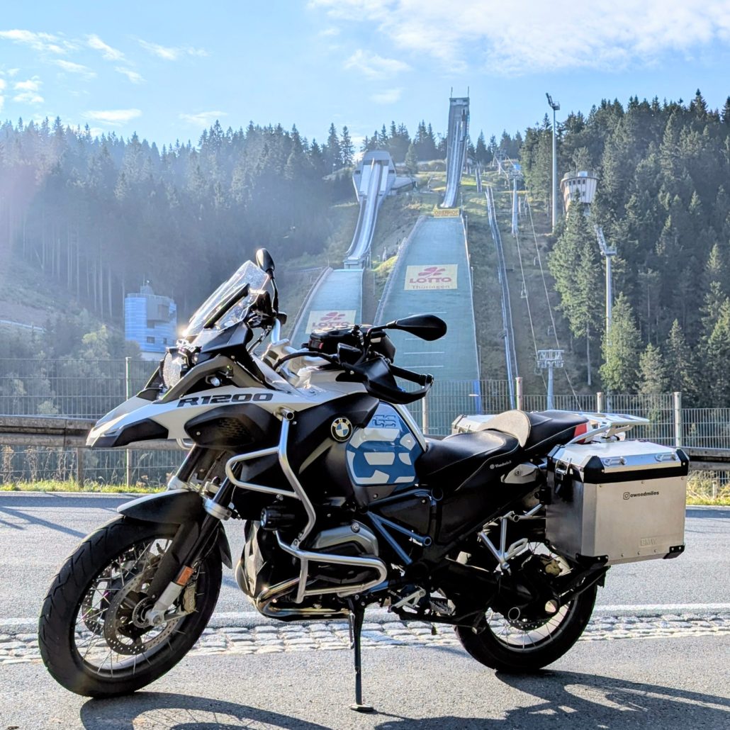 Weiße BMW R 1200 GS mit silbernen Alukoffern parkt auf Asphalt vor einer großen Skisprungschanze im Wald.