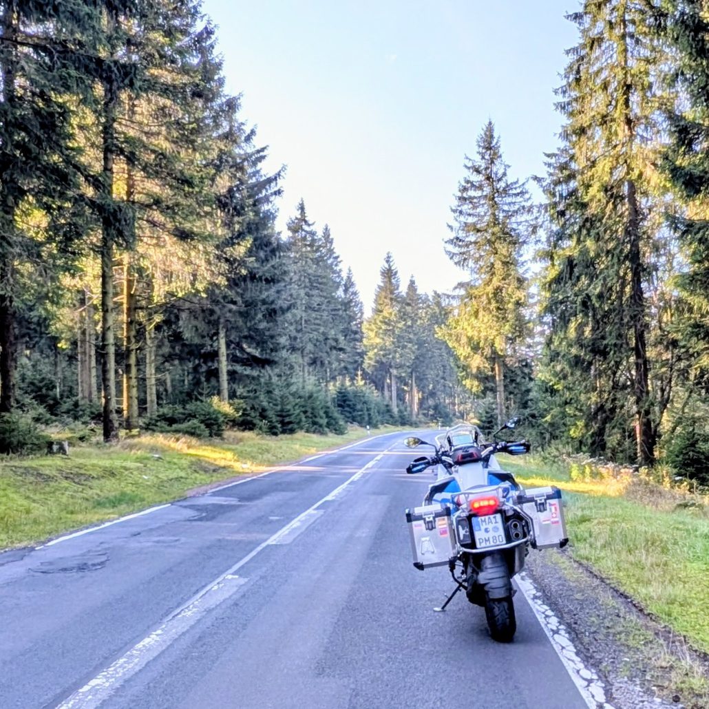 Heckansicht einer weißen BMW R 1200 GS Adventure mit silbernen Alukoffern auf einer asphaltierten Waldstraße im Sonnenlicht.