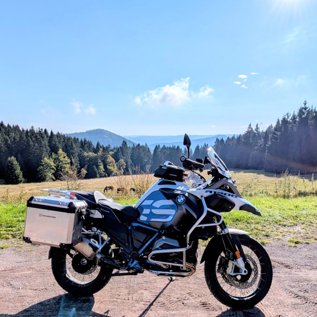 Weiße BMW R 1200 GS Adventure mit Alukoffern und Tanktasche steht auf unbefestigtem Weg vor einer Bergwiesenlandschaft.