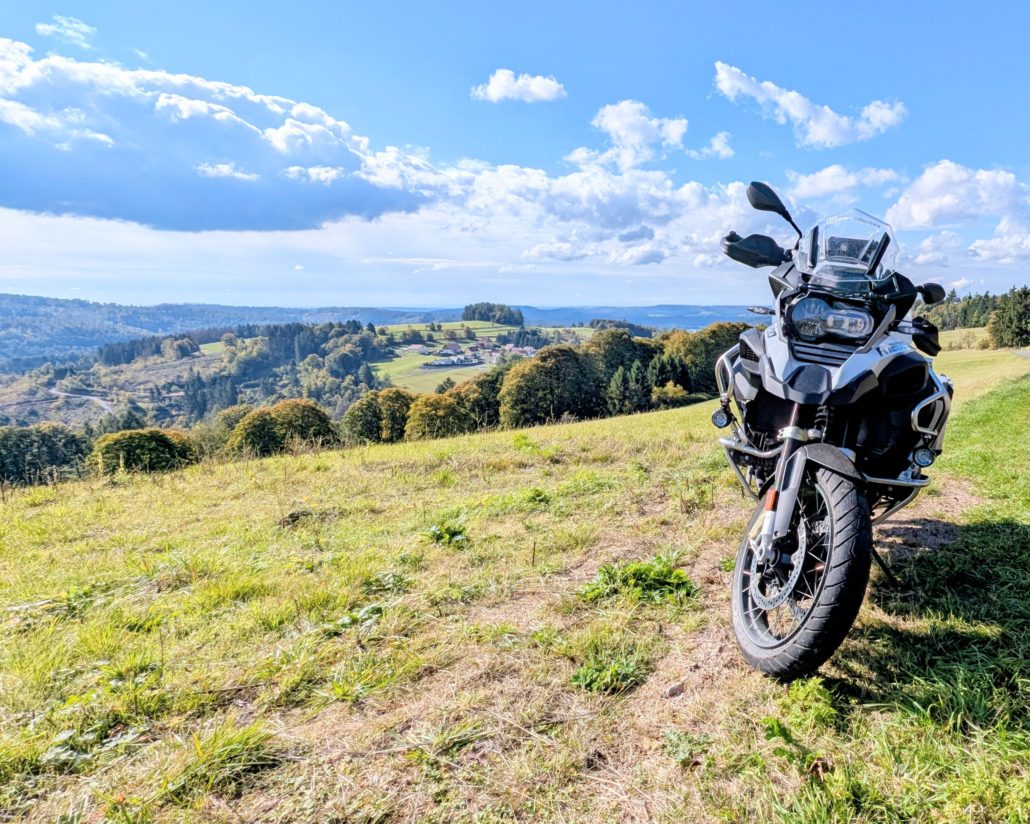 Silberne BMW R 1200 GS Adventure steht auf einer Wiese mit Blick auf bewaldete Hügel und weite Landschaft unter blauem Himmel.