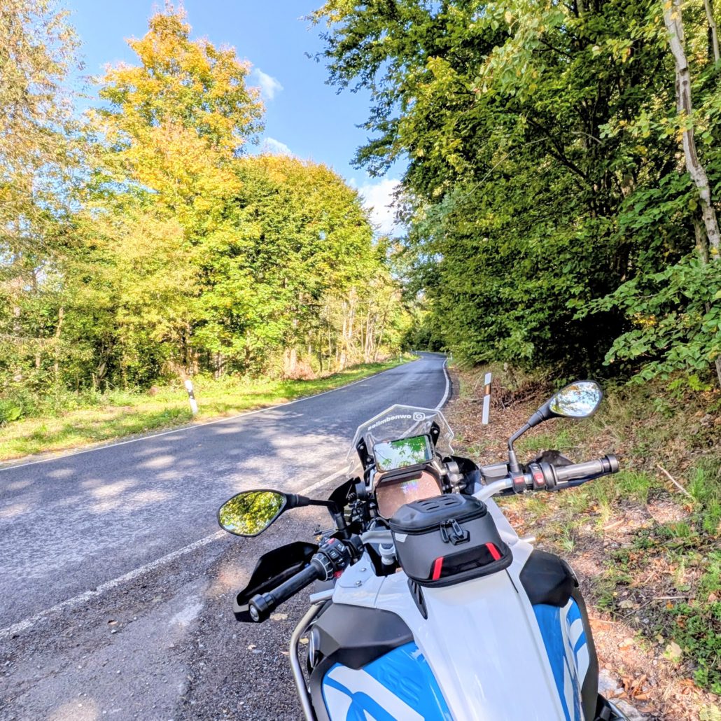 Cockpit einer weißen BMW R 1200 GS mit Navigationssystem und Tanktasche am Rand einer asphaltierten Straße im Wald.