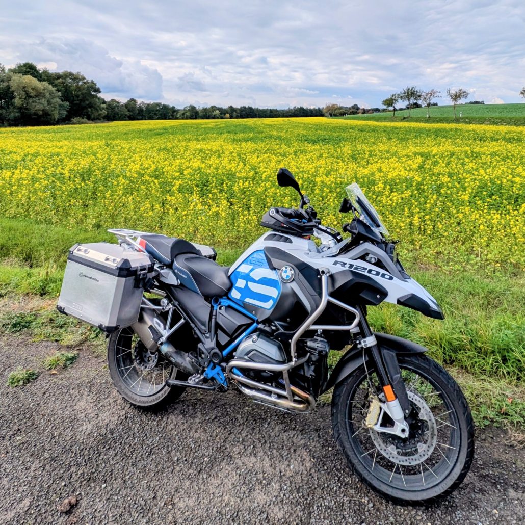 BMW R 1200 GS Adventure Motorrad, weiß-blau, mit Aluminium-Seitenkoffern auf Schotterweg vor einem leuchtend gelben Rapsfeld.