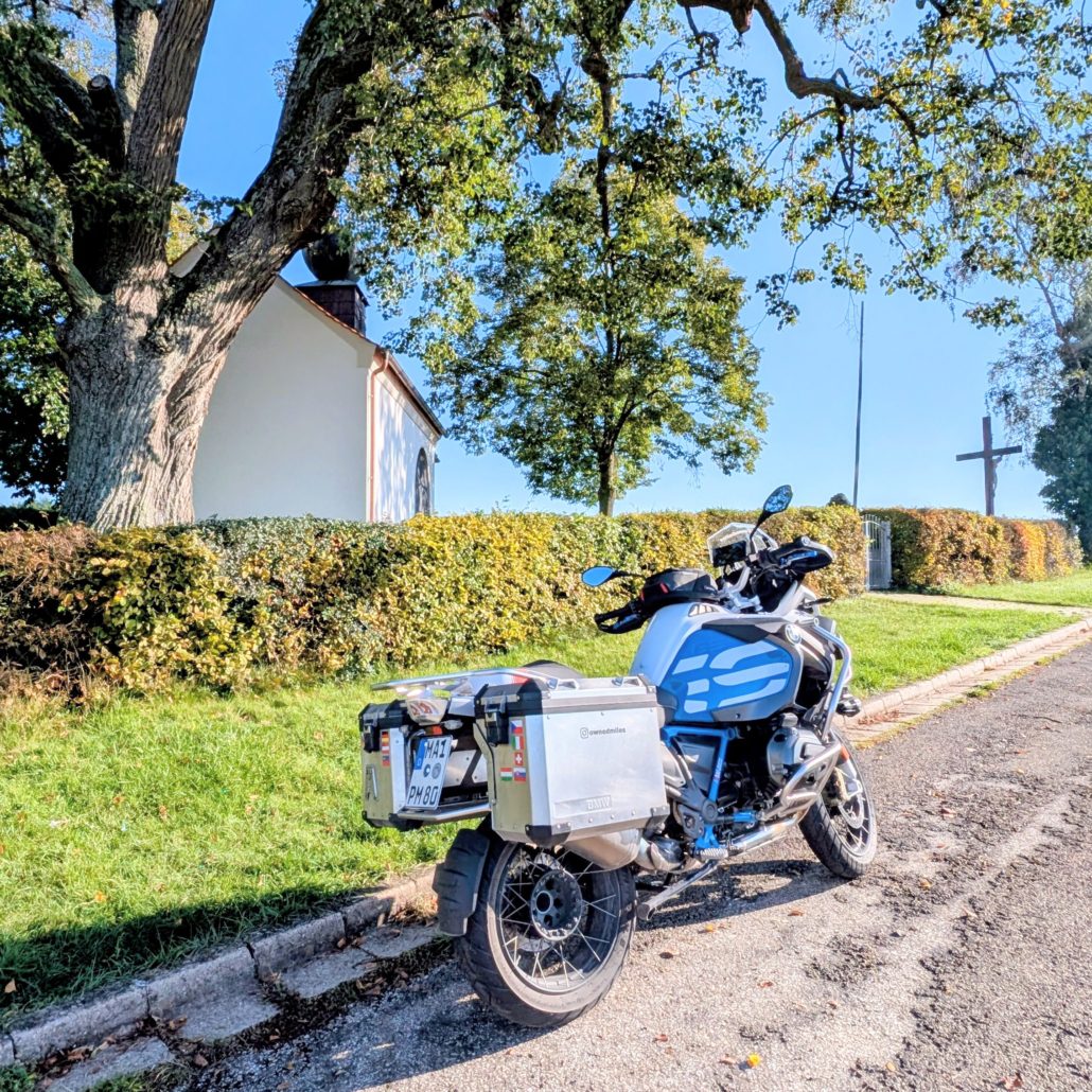 Weiße BMW R 1200 GS Adventure mit blauem Dekor und Alukoffern parkt am Straßenrand vor einer kleinen Kapelle und einem Holzkreuz.