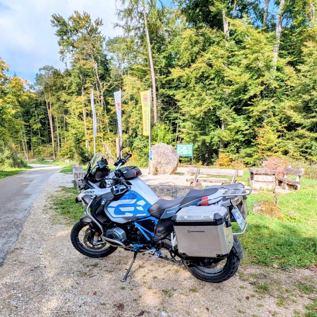 Weiße BMW R 1200 GS Adventure mit blauem Dekor und silbernen Aluminium-Koffern parkt auf Schotter vor Waldrand.