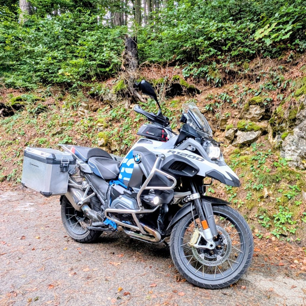 BMW R 1200 GS Motorrad in Weiß und Blau, bepackt mit schwarzen und silbernen Taschen, auf einem Waldweg in der Totalen.