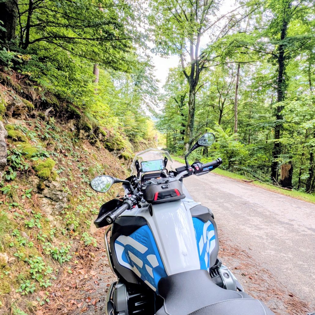 BMW R 1200 GS Motorrad in Weiß und Blau, bepackt mit schwarzen und silbernen Taschen, auf einem Waldweg in der Totalen.