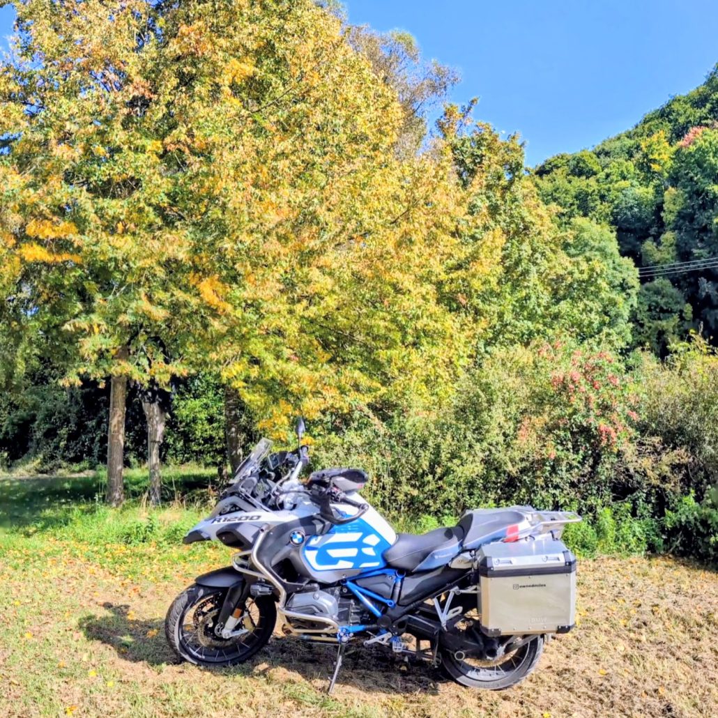 BMW R 1200 GS Adventure Motorrad, weiß/blau, mit silbernen Alukoffern, steht auf einer Lichtung vor herbstlich gefärbten Bäumen.