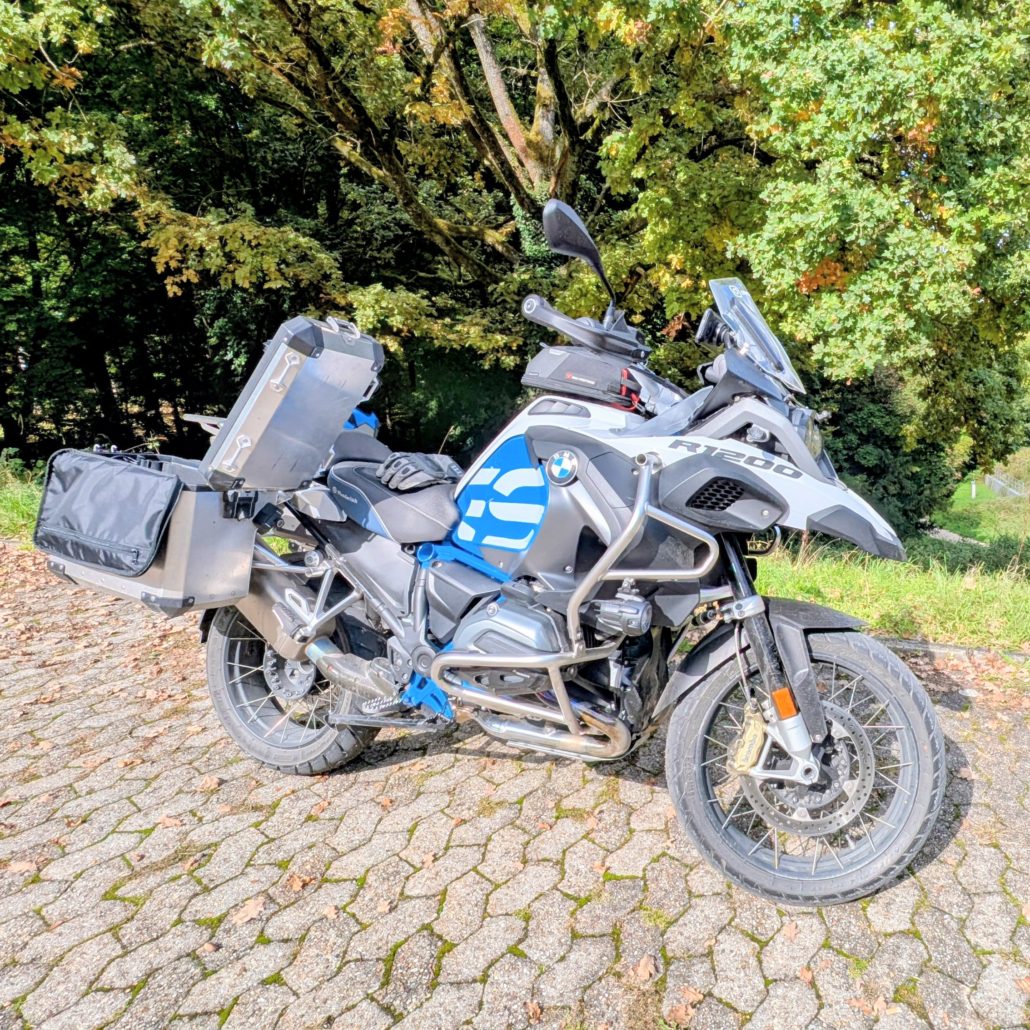 Weiße BMW R 1200 GS mit blauen Akzenten, Gepäckkoffern aus Aluminium und schwarzer Tasche steht auf Kopfsteinpflaster.