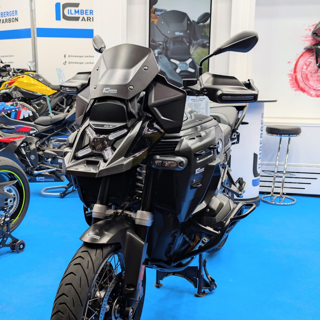 Schwarze BMW R 1300 GS Adventure mit Carbon-Teilen am Messestand von Ilmberger Carbon auf blauem Teppichboden.