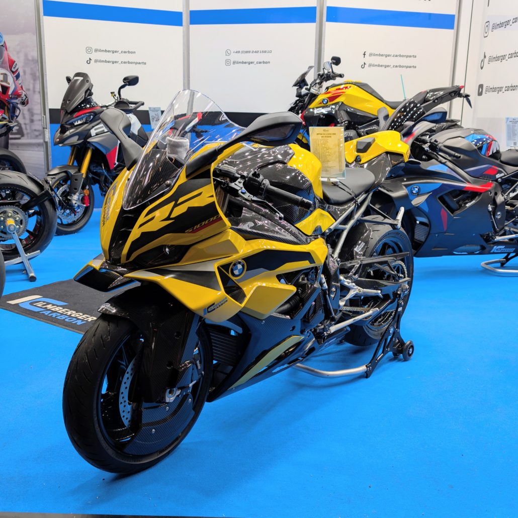 Gelbe BMW S 1000 RR mit Carbon-Verkleidung von Ilmberger Carbon auf blauem Messeboden vor weiteren Motorrädern.