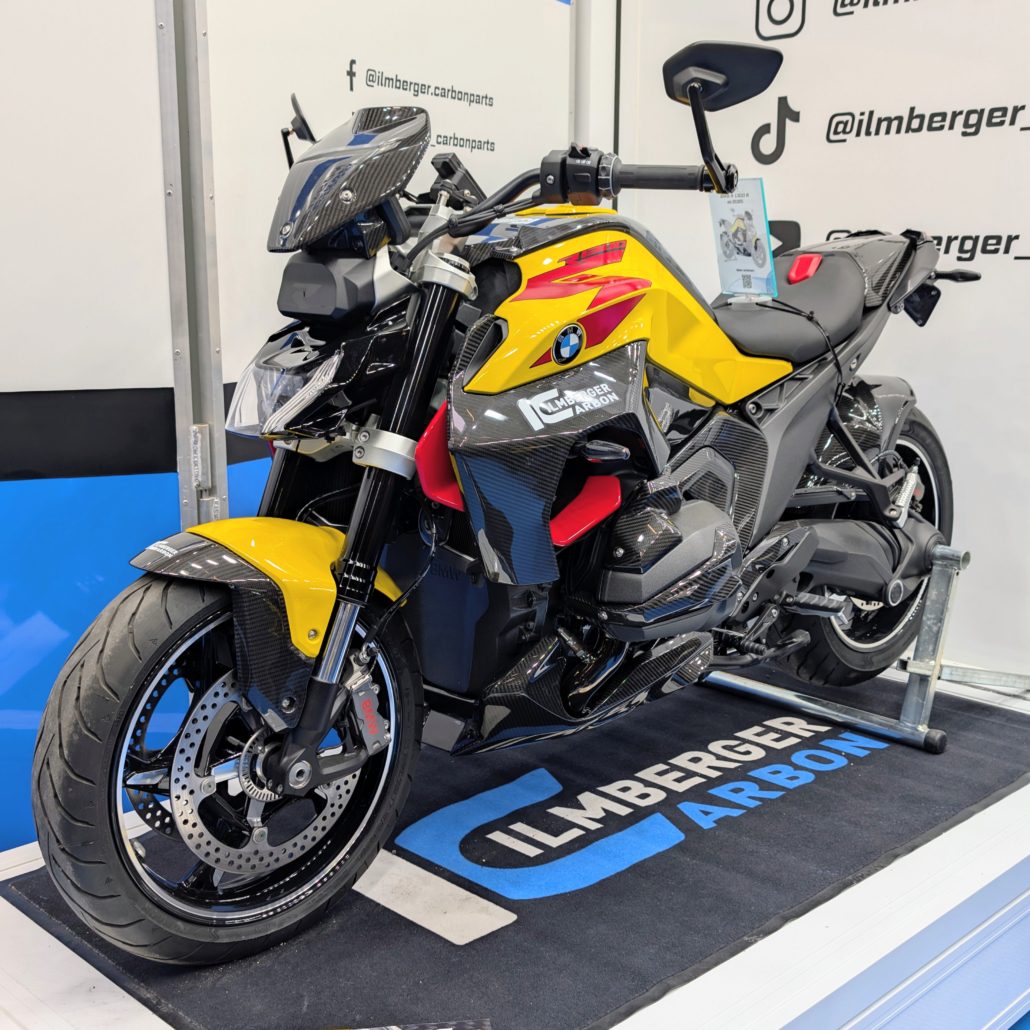 Gelbe BMW R 1200 R mit Carbon-Teilen von Ilmberger Carbon auf einem Messestand mit Marken-Teppich und Social-Media-Logos.