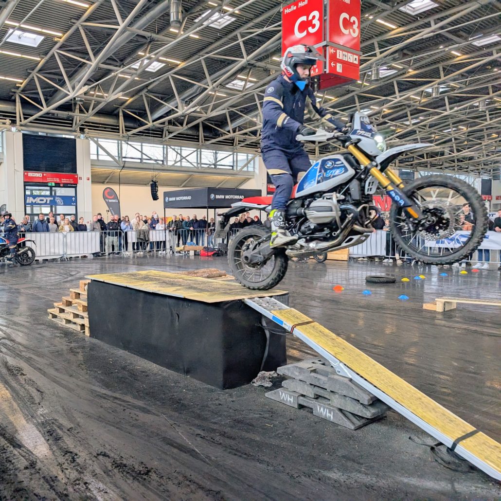 Motorradfahrer springt mit einer BMW R12 G/S über eine Rampe während einer Stuntshow in der Messehalle C3 der IMOT.