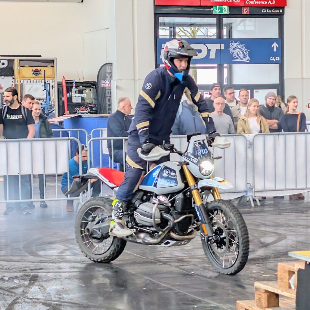 Motorradfahrer mit BMW-Ausrüstung führt Stunt auf einer BMW R 12 G/S vor Publikum in einer Messehalle vor.