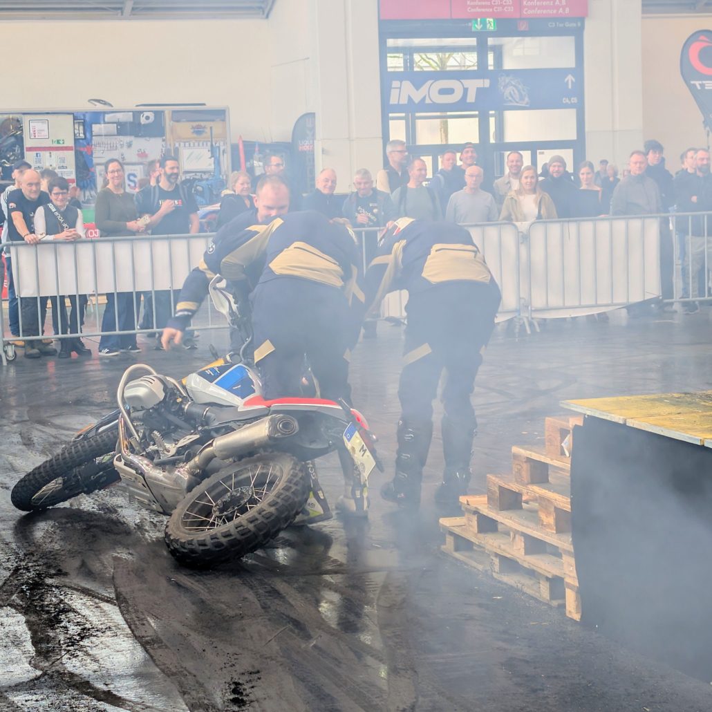 Zwei Helfer richten ein gestürztes BMW Motorrad nach einem Stunt auf der IMOT Messe im dichten Nebel vor Zuschauern auf.