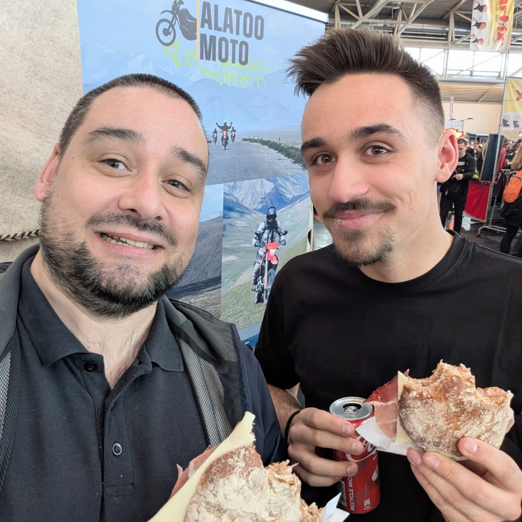 Zwei lächelnde Männer essen belegte Brötchen vor einem Alatoo Moto Banner auf einer Messe.