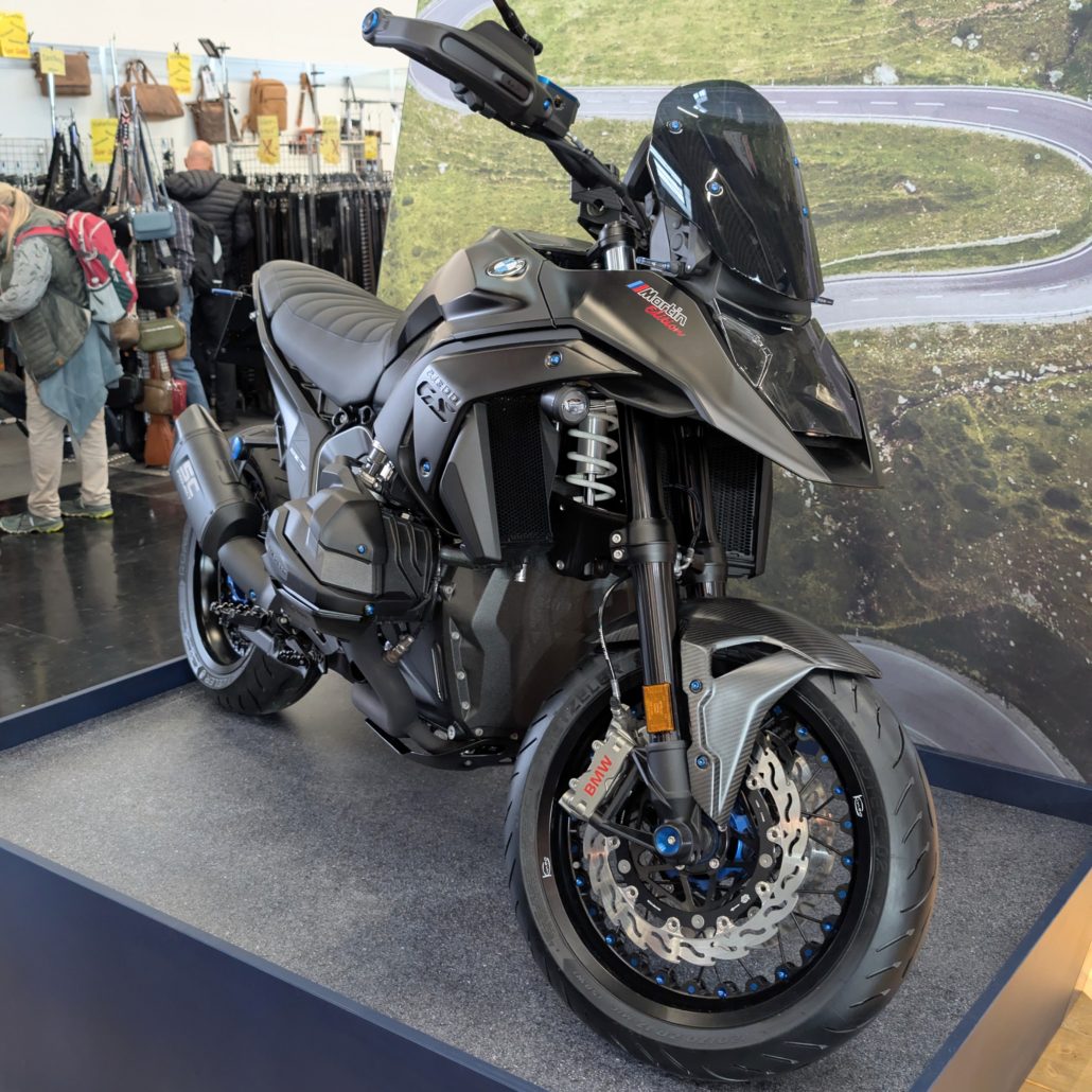 Mattschwarze BMW R 1300 GS Martin Edition mit Carbon-Elementen und blauen Akzenten auf einem Podest bei einer Messe.