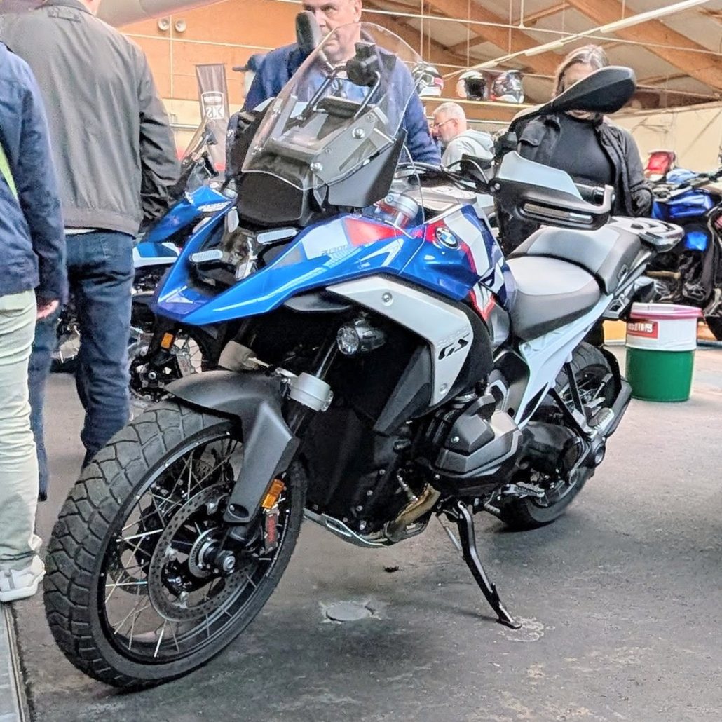 Blau-weiße BMW R 1300 GS Adventure mit Speichenrädern und GS-Schriftzug auf einer Messe mit Besuchern im Hintergrund.