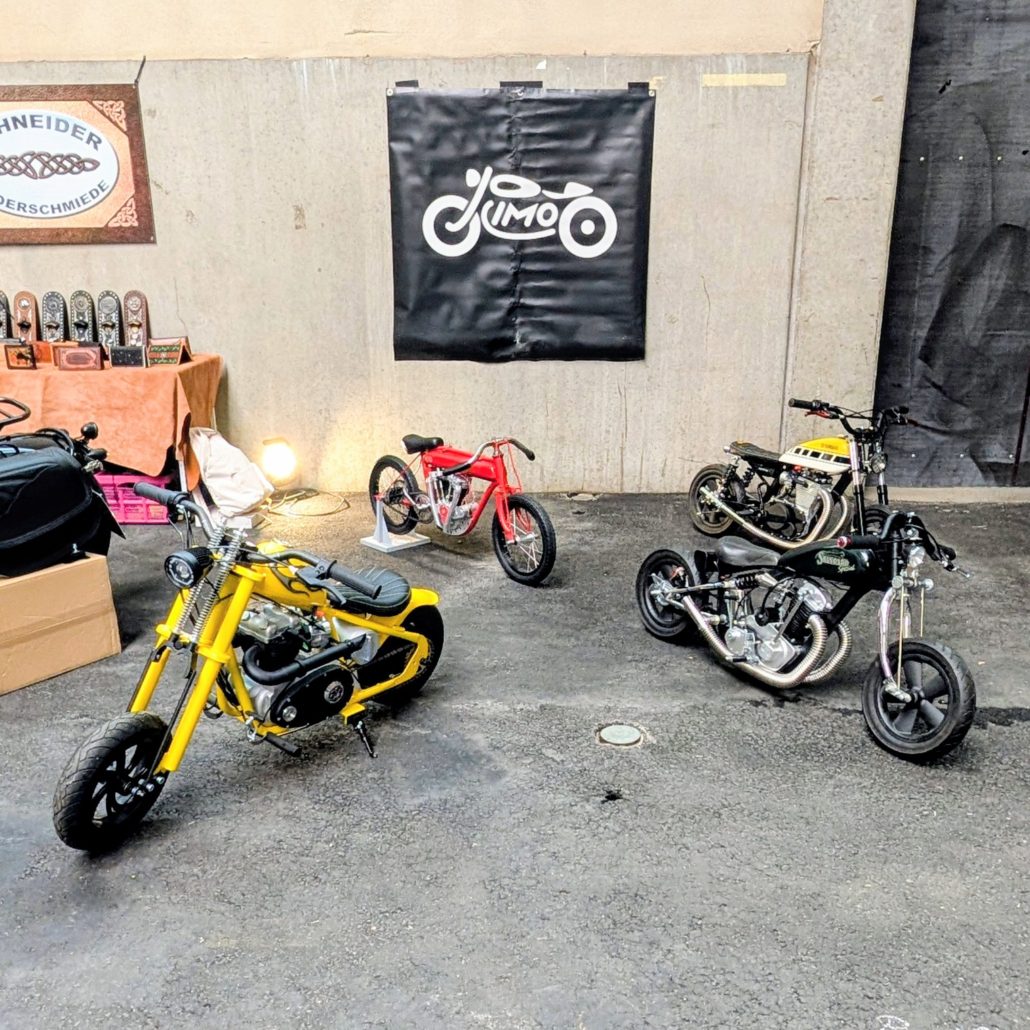 Vier Mini-Motorräder in Gelb, Rot, Schwarz und Weiß vor einer Betonwand mit schwarzem JUMO-Banner und Werkstatt-Deko.