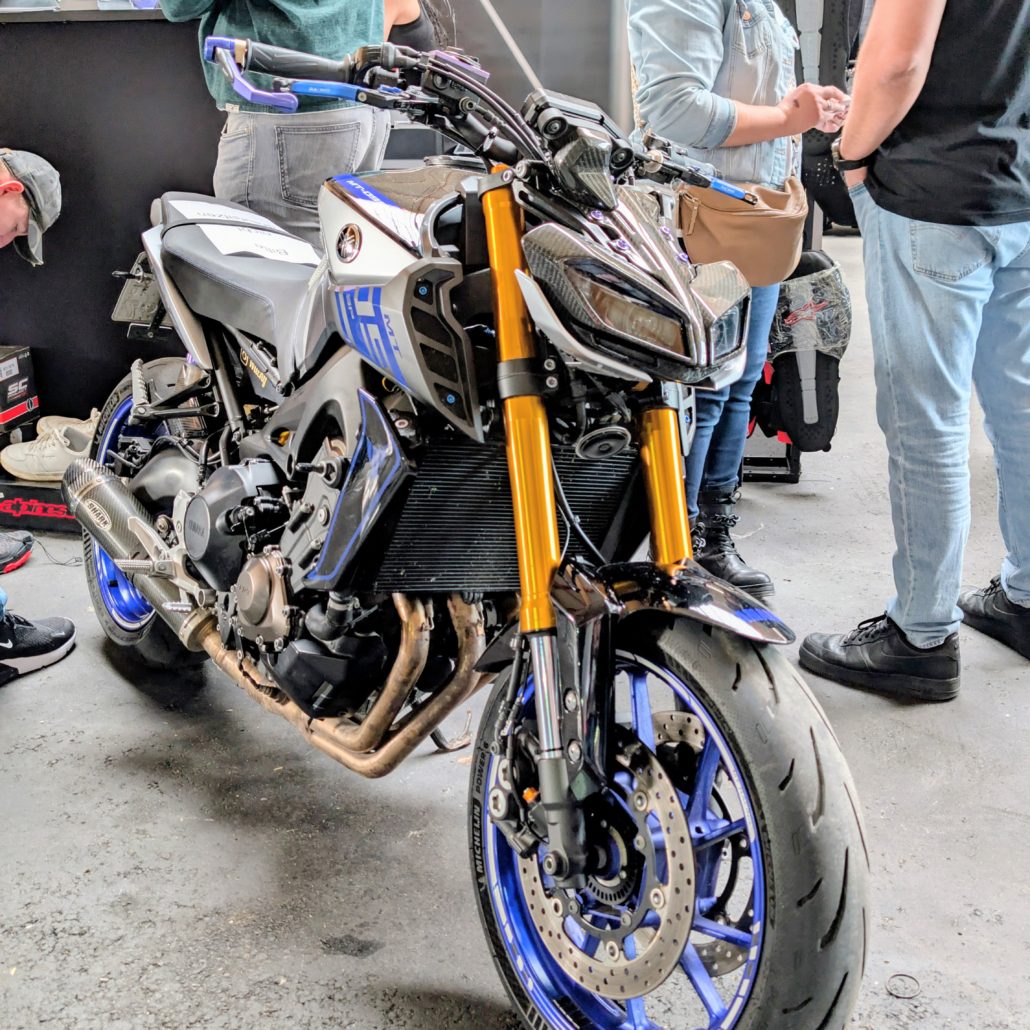 Silber-blaue Yamaha MT-09 mit goldener Gabel, blauen Felgen und Shark-Auspuff auf einer Messe mit Besuchern im Hintergrund.