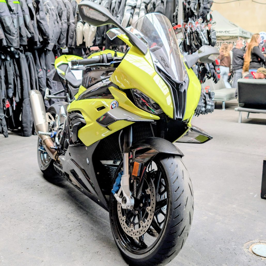Gelbe BMW S 1000 RR mit Carbon-Frontfender und Winglets vor einer Wand mit Motorradbekleidung auf einer Messe.