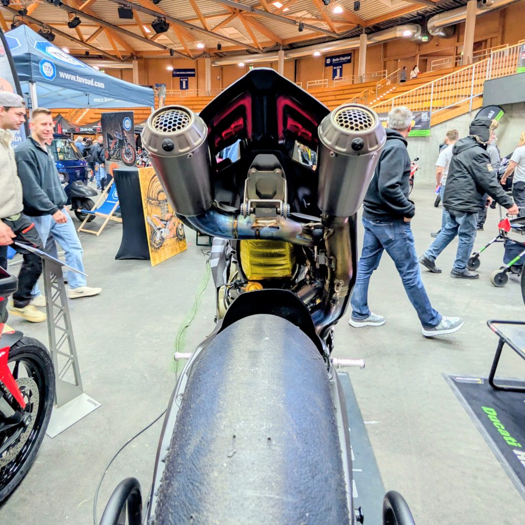 Heckansicht eines Rennmotorrads mit breitem Slick-Reifen und markanter Underseat-Doppelauspuffanlage auf einer Messe.