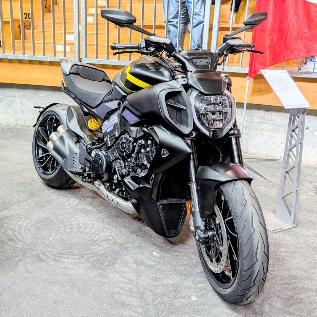 Schwarze Ducati Diavel V4 mit gelben Akzenten, charakteristischem LED-Scheinwerfer und Vierfach-Auspuff in einer Ausstellung.