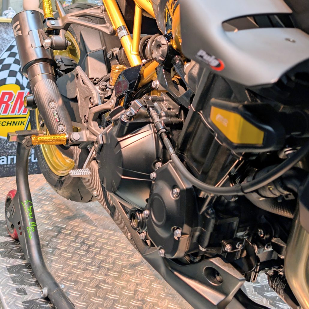 Detailaufnahme eines Motorradmotors mit goldenem Rahmen, Carbon-Auspuff und Puig-Komponenten auf einem Montageständer.