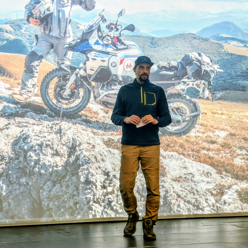 Sprecher in blauem Pullover und brauner Hose vor Leinwand mit BMW-Motorrad in Berglandschaft.