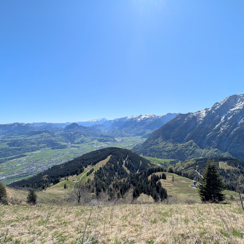 Panorama-Blick von der Rossfeld Panoramastraße über schneebedeckte Alpengipfel und grüne Täler bei strahlendem Sonnenschein.